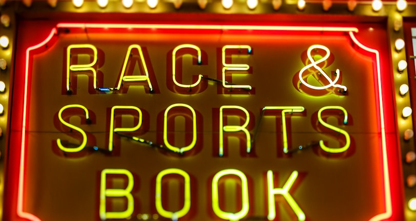 Bright neon lights on a marquee sign display the words 'RACE & SPORTS BOOK'. The sign is surrounded by rows of yellow and white lights against a red background, evoking a vibrant, energetic atmosphere typically associated with casinos or entertainment venues.