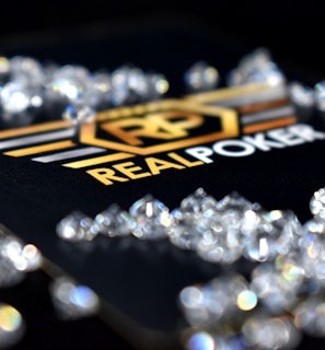 A collection of sparkling, clear gemstones surrounds a dark surface featuring the prominent logo and text 'Real Poker.' The logo design includes gold and white elements set against a black background.